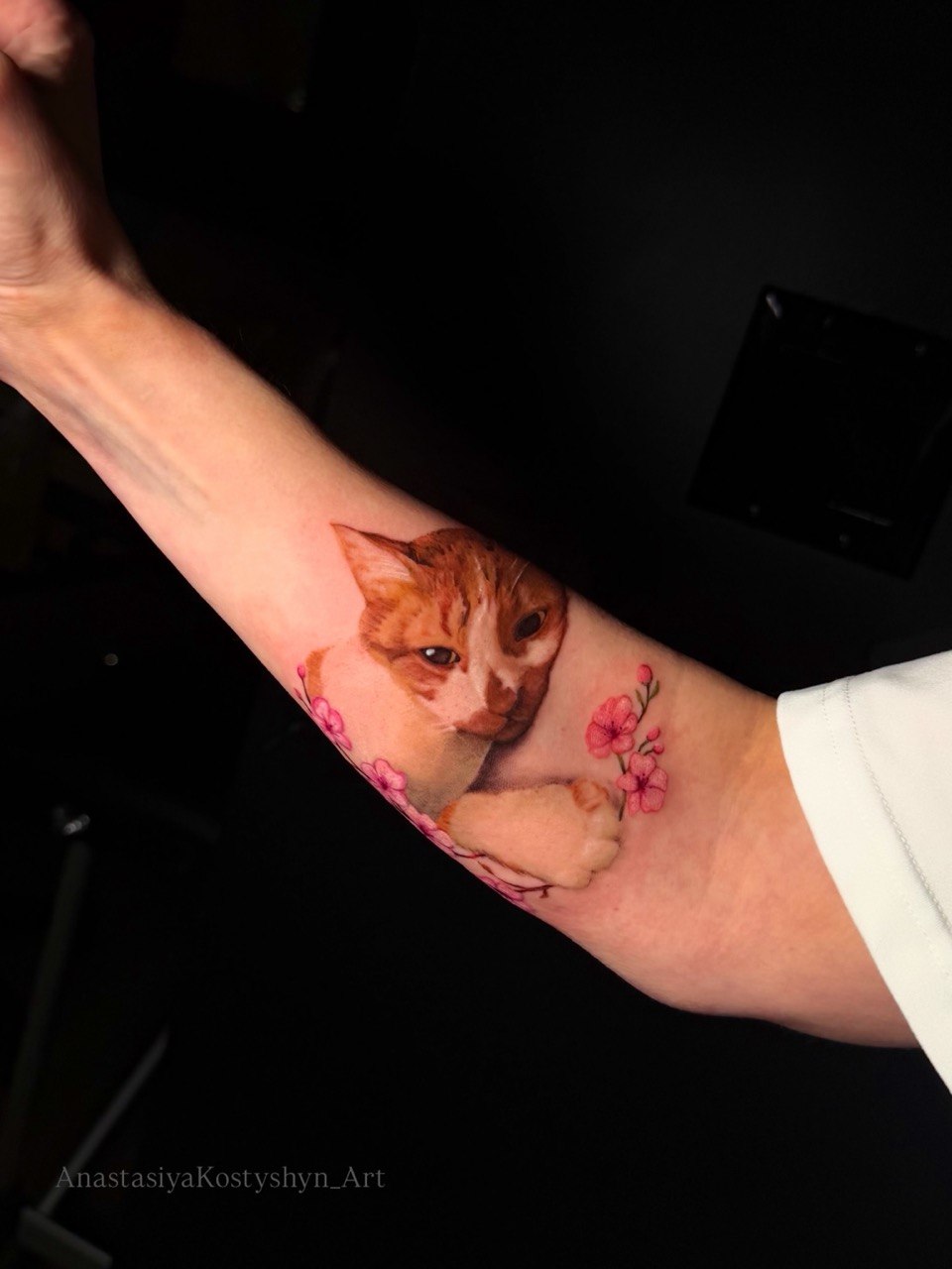 Realistic color cat portrait tattoo on the forearm accented with soft pink cherry blossom flowers